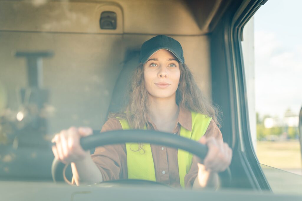woman with a trucker hat on and a yellow vest driving her transfer truck